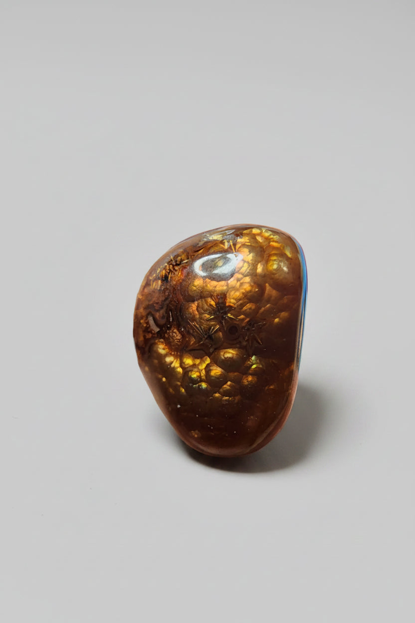 A close-up of a polished brown gemstone or resin cabochon held between fingers. The stone has a glossy, rounded teardrop shape with a rich amber-brown color and internal crystalline or metallic gold patterns.