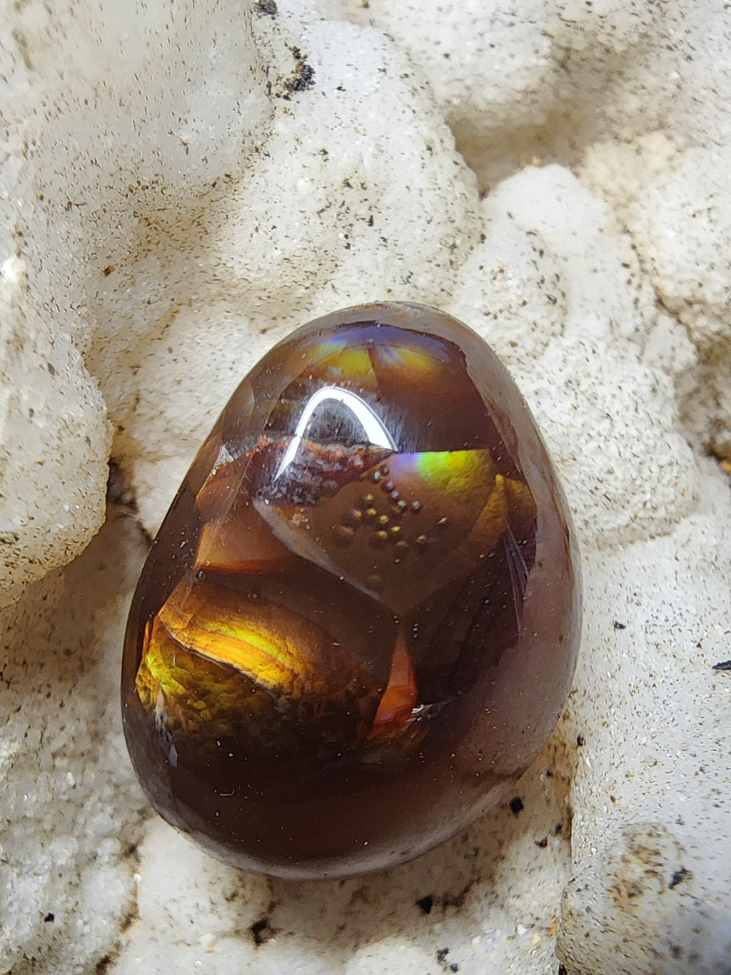 A polished, smooth oval gemstone with a rich amber-brown color and iridescent yellow-green internal reflections, resting on a textured white crystal cluster.