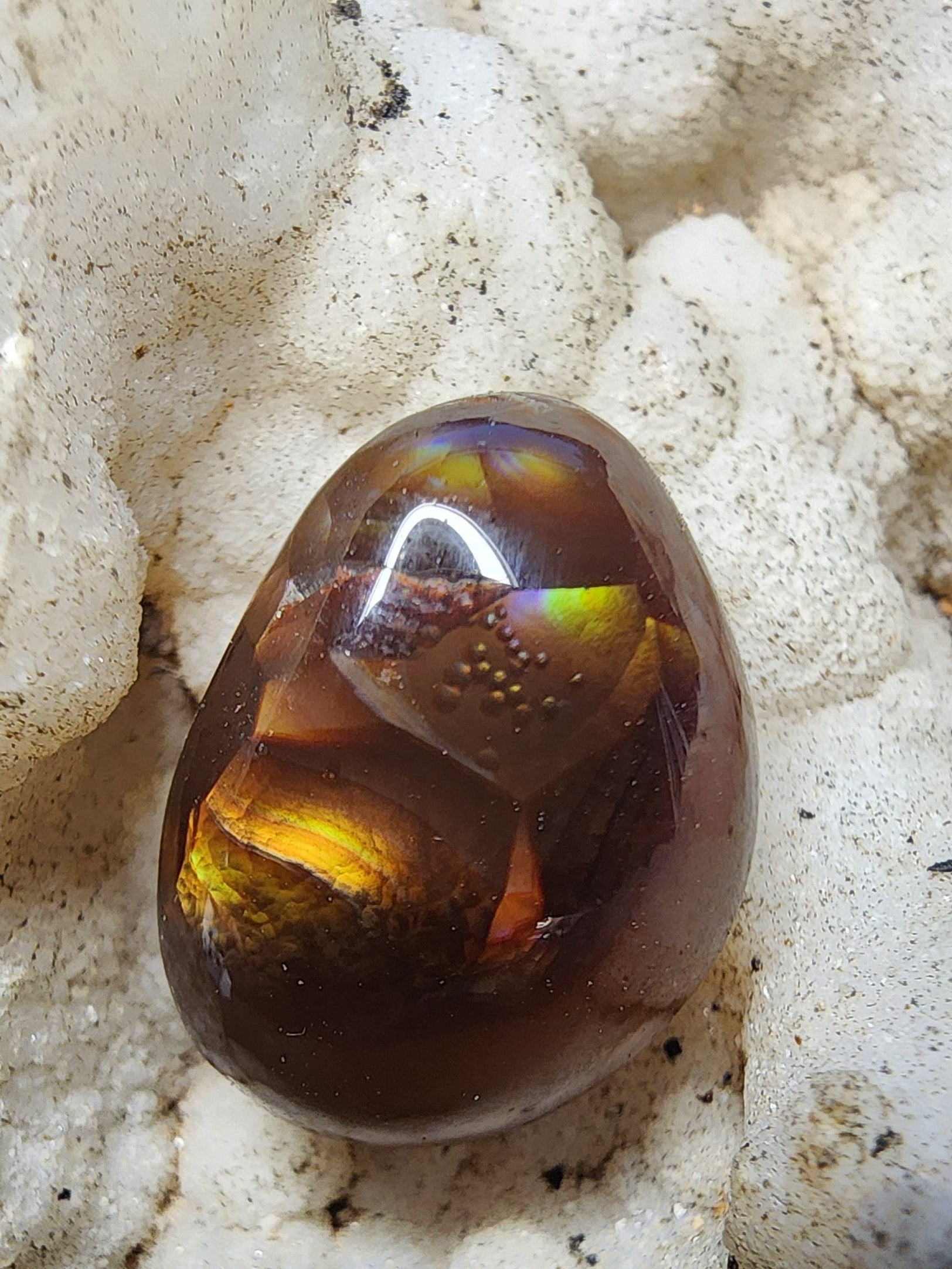 A polished, smooth oval gemstone with a rich amber-brown color and iridescent yellow-green internal reflections, resting on a textured white crystal cluster.