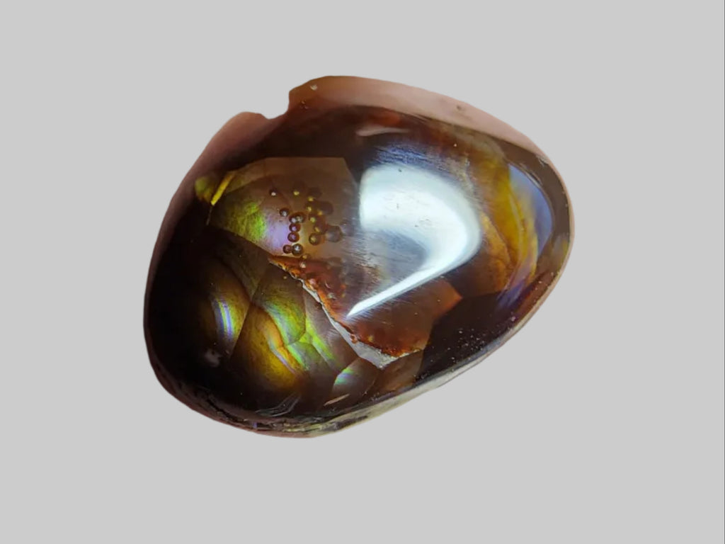 A polished brown gemstone with golden-yellow inclusions, resting on a dark gray textured surface next to a centimeter ruler for scale.
